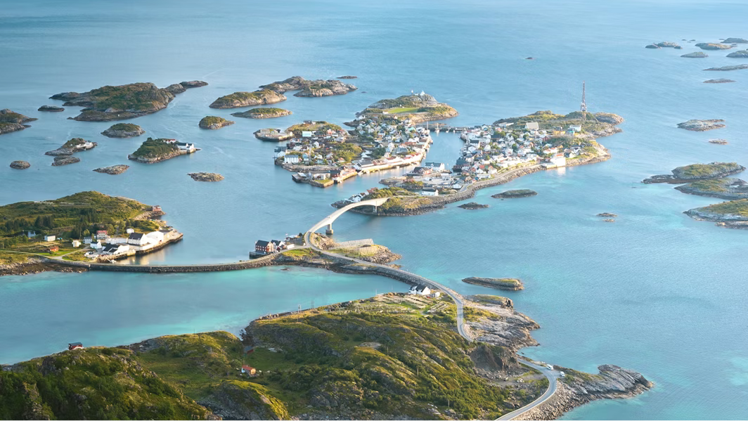Aerial view of a coastal archipelago with turquoise-blue water, small rocky green islands, and a connected village of white and colorful houses linked by winding roads and bridges stretching across the sea.