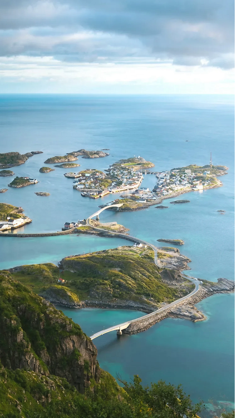 Aerial view of a coastal archipelago with turquoise-blue water, small rocky green islands, and a connected village of white and colorful houses linked by winding roads and bridges stretching across the sea.